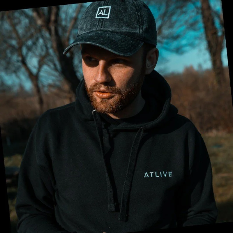 A young man with a beard wearing a black hoodie with "ATLIVE" on it and a black cap with a logo, outdoors with trees in the background.