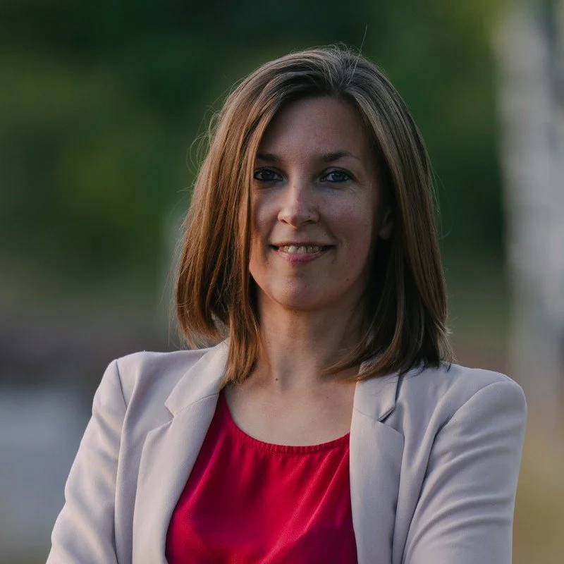 A woman with shoulder-length brown hair, smiling, outdoors with blurred green trees in the background. She is wearing a light-colored blazer and a red top.