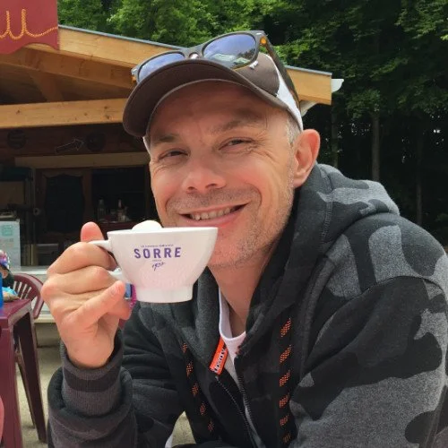 Smiling man wearing a cap and sunglasses on his head, holding a small cup of coffee outdoors at a wooden cafe with trees in the background.