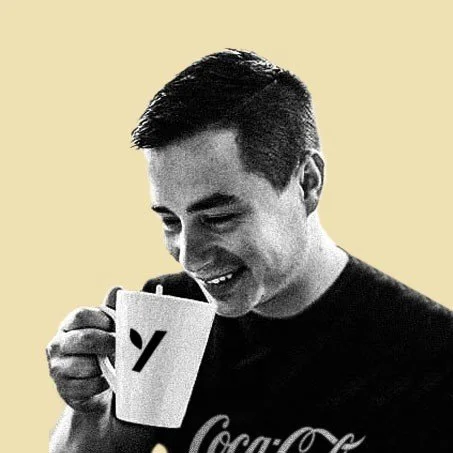 A smiling young man holding a white mug with a logo, wearing a black shirt, against a plain light background.