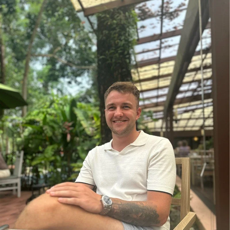 A smiling man with short brown hair and a tattooed arm sitting at a table outdoors in a lush green setting