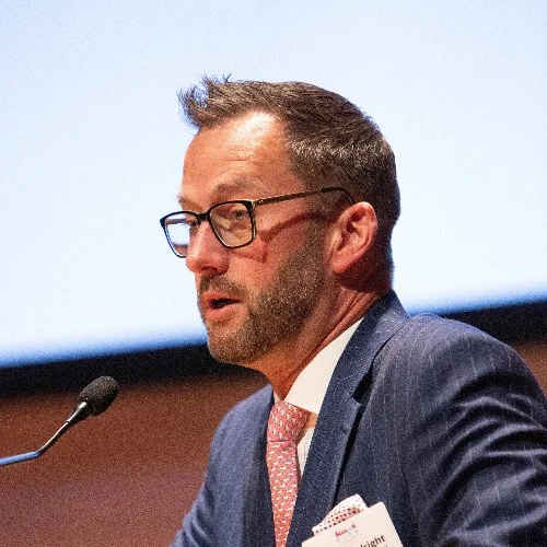 A man wearing glasses and a suit speaking at a podium during a conference or presentation.