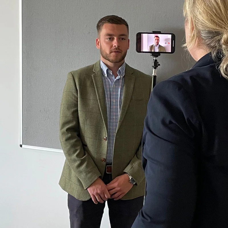 A young man is being recorded on a smartphone for a video or interview, standing against a plain gray wall in an indoor setting.