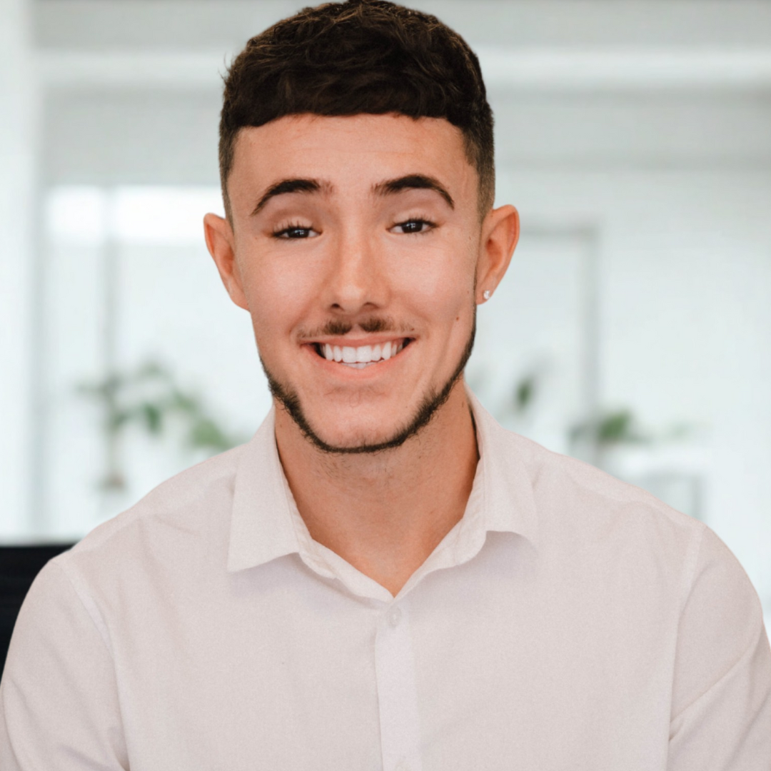 Young man with short dark hair, a light beard, and earrings, smiling in an office setting with a light background and potted plants.