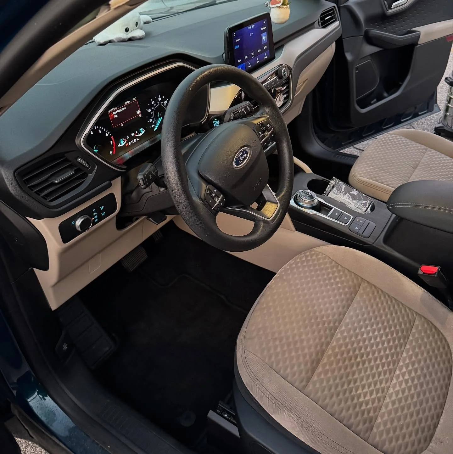 Interior of a Ford vehicle showing the steering wheel, dashboard with instrument cluster, central touchscreen display, beige and black seats, and center console.