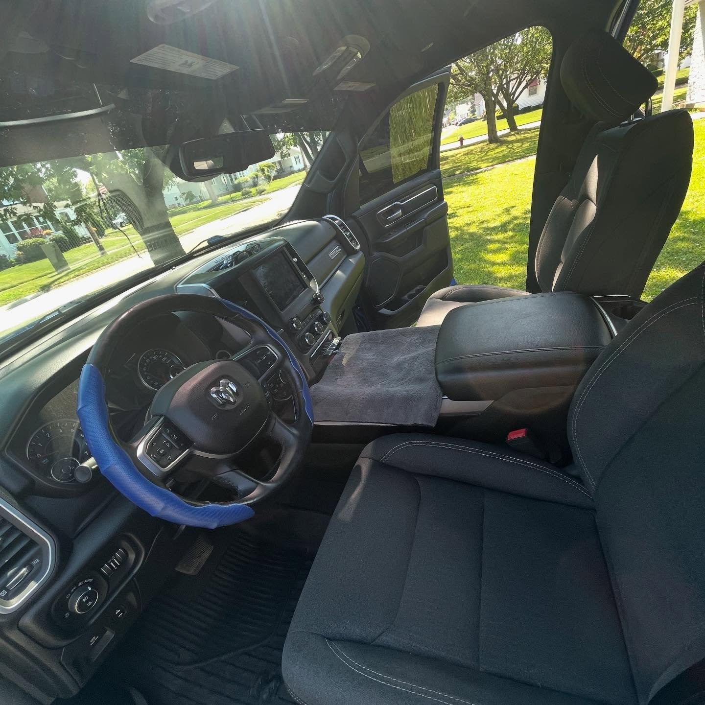 The interior of a vehicle showing the driver's seat, steering wheel with blue cover, dashboard, and passenger seat with a fold-down armrest.