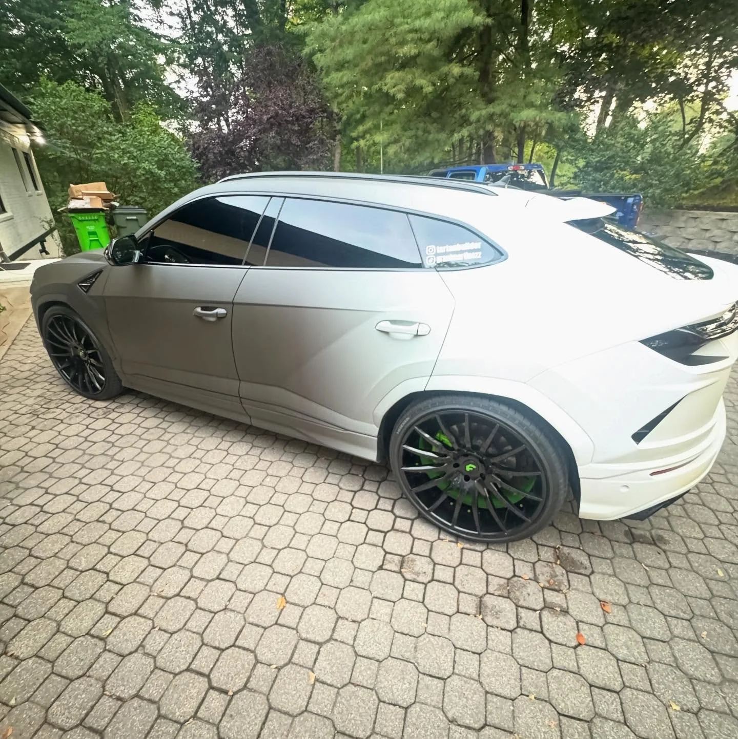 A white electric SUV with black tinted windows and black custom wheels parked on a stone pavement in a backyard with trees and greenery in the background.