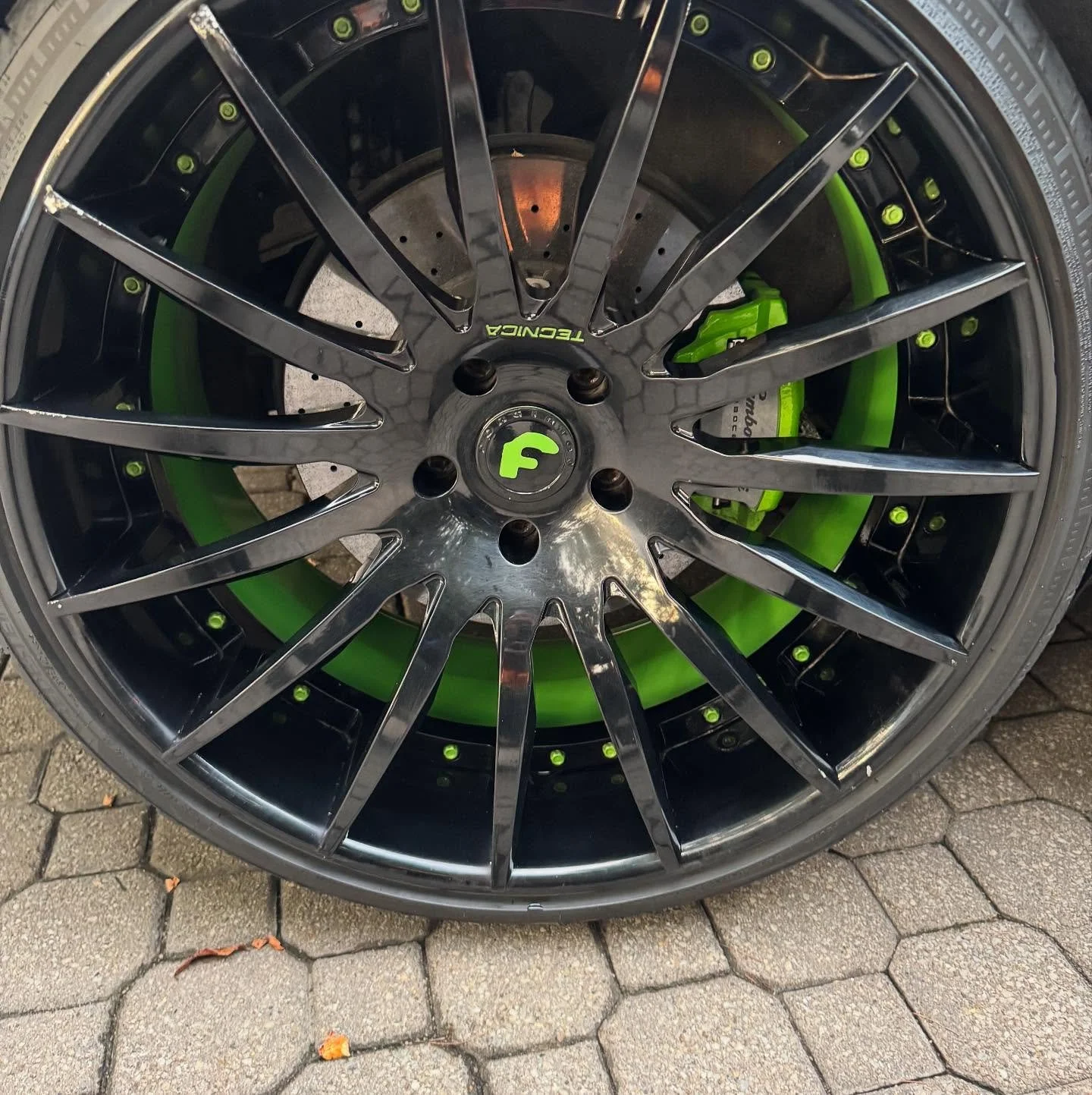 Close-up of a black car wheel with green accents on the brake caliper and rim, with a multi-spoke rim design, on a paved surface.