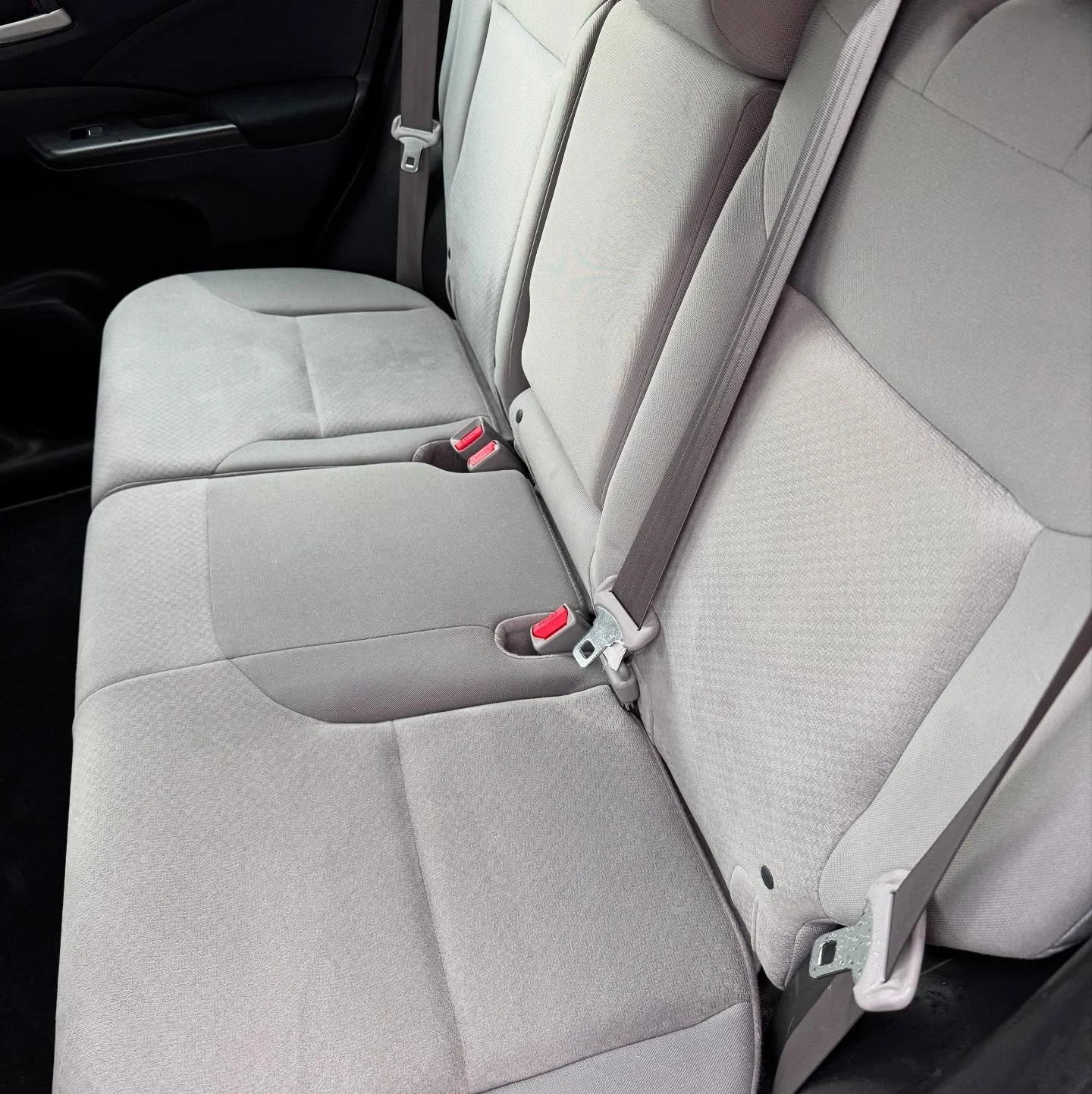 Inside of a vehicle showing the beige fabric backseat with three seat belts and seatbelt buckles.