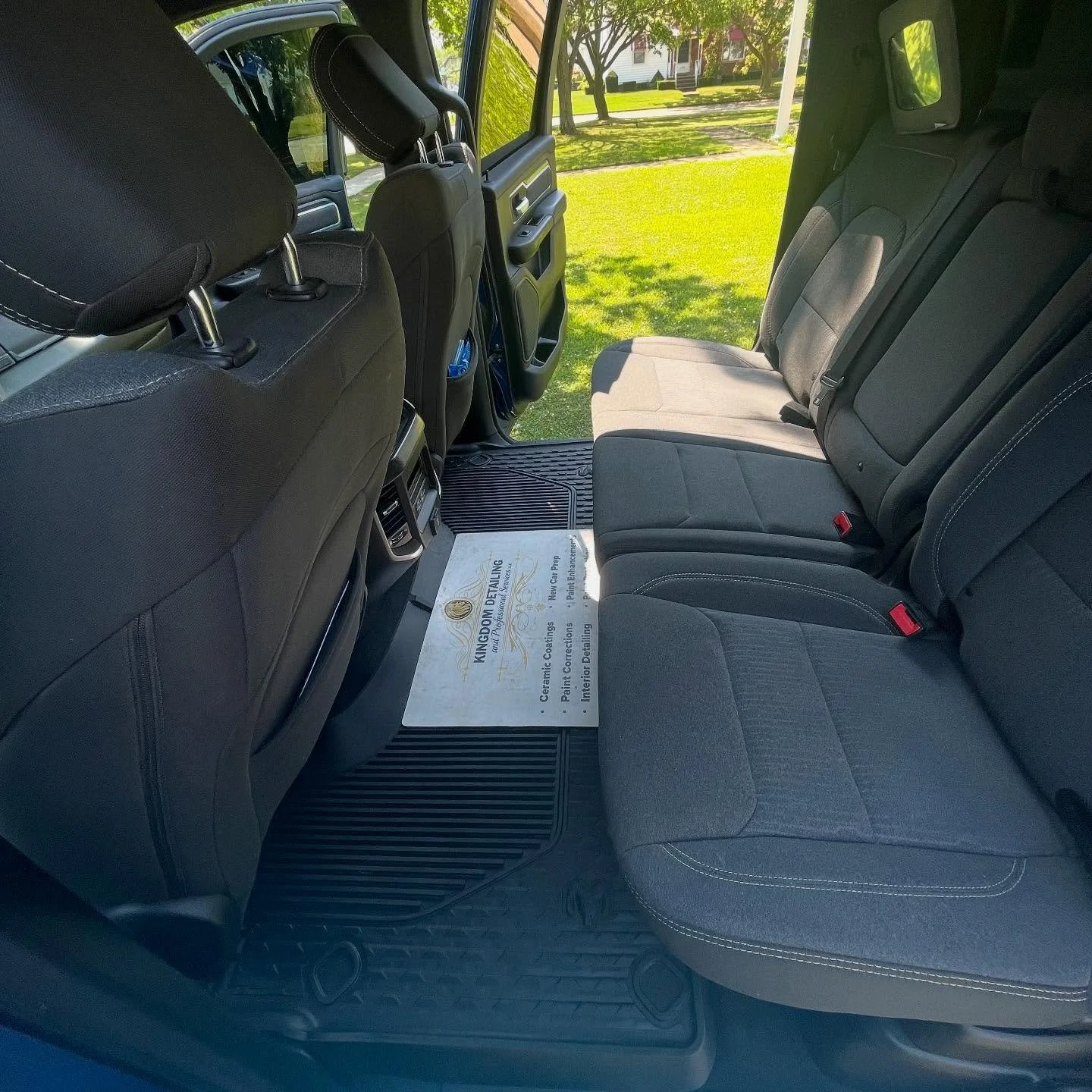 The backseat interior of a vehicle, showing three black fabric seats, a door on the right side, and a view of green grass and trees outside through the windows.