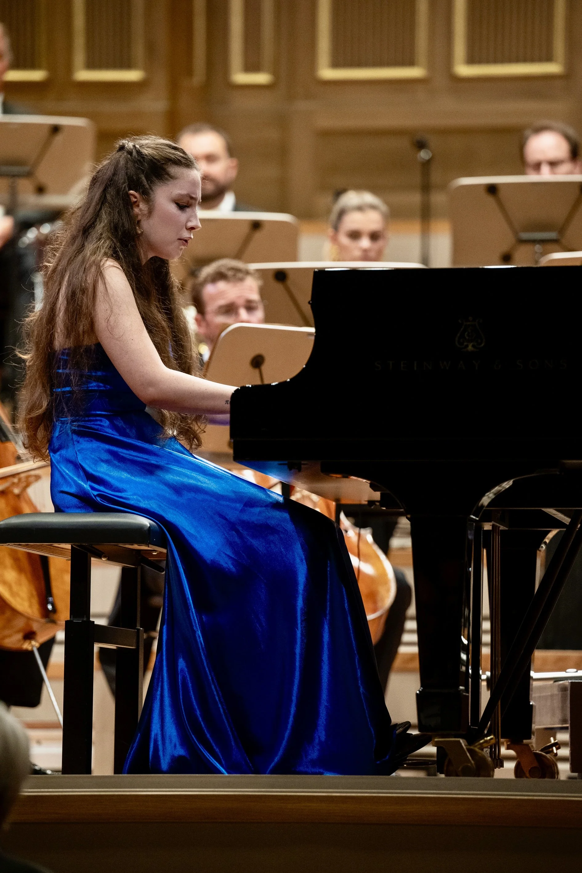 Una giovane donna con capelli lunghi e ricci suona un pianoforte Steinway & Sons, indossa un abito blu in un concerto con un'orchestra sullo sfondo.