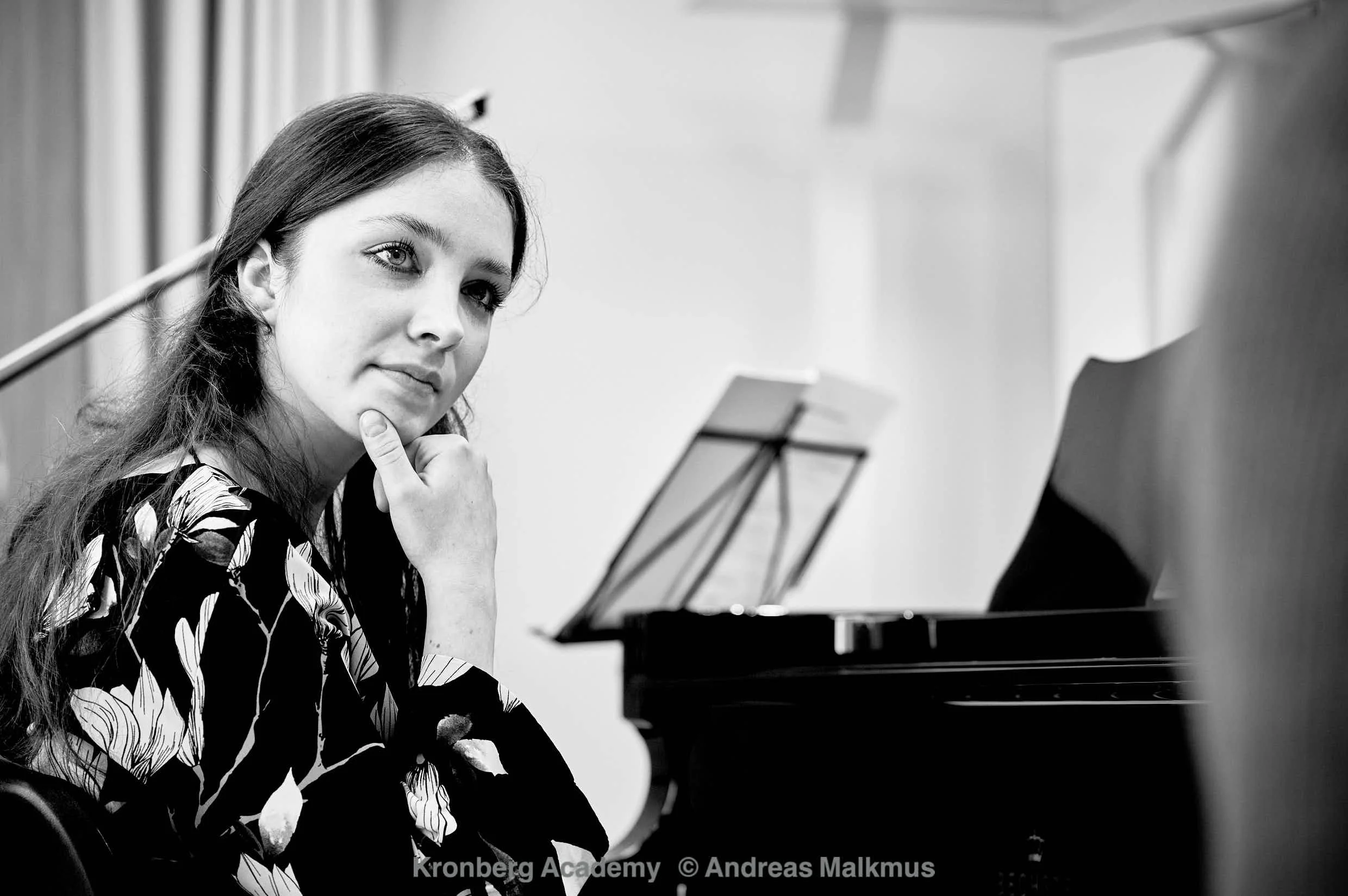 Ragazza con capelli lunghi e occhi chiari seduta davanti a un pianoforte, con il mento appoggiato sulla mano e uno sguardo pensieroso.