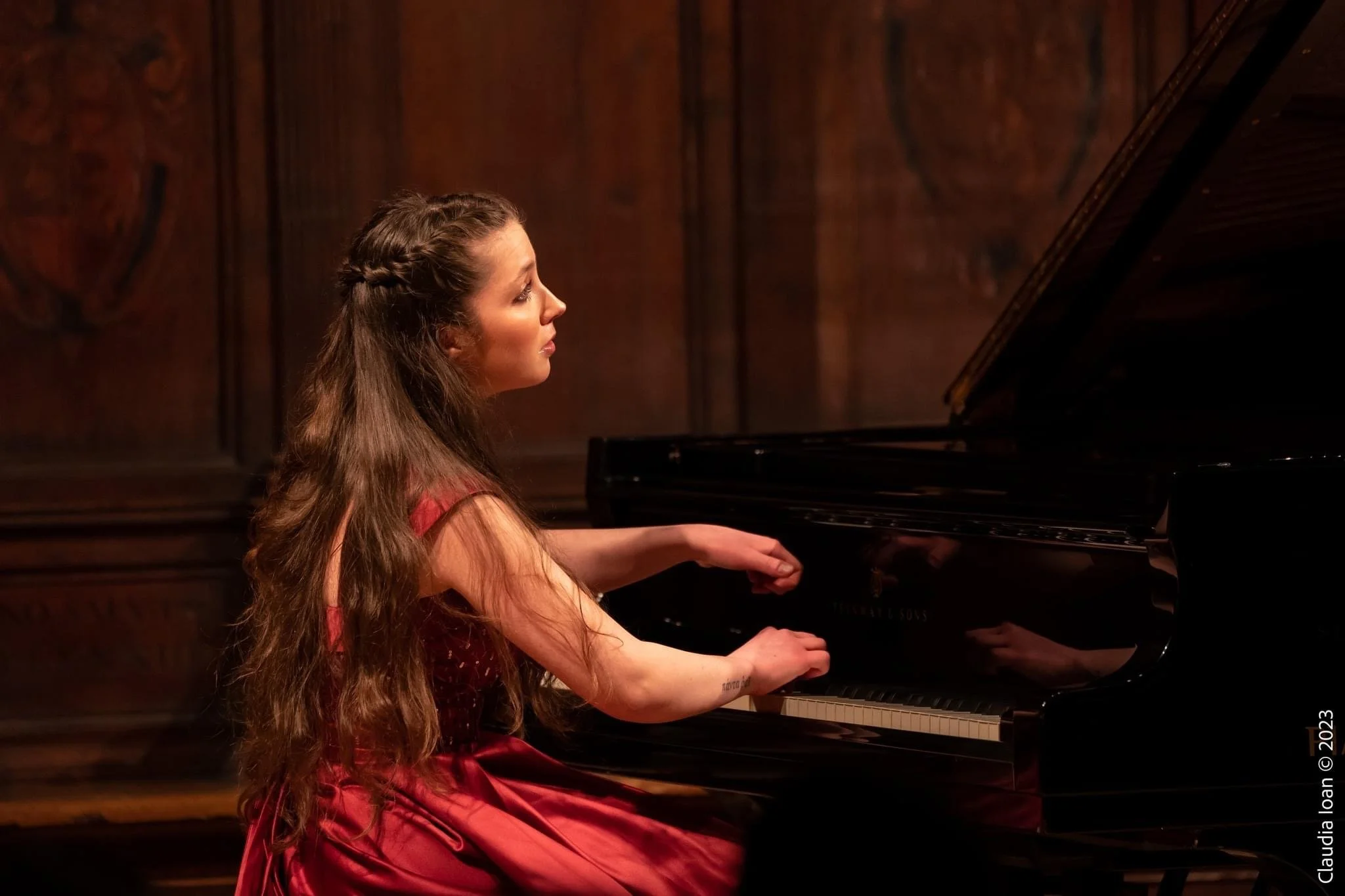Una giovane donna seduta davanti a un pianoforte, che sembra concentrata mentre suona, con uno sfondo di pannelli di legno
