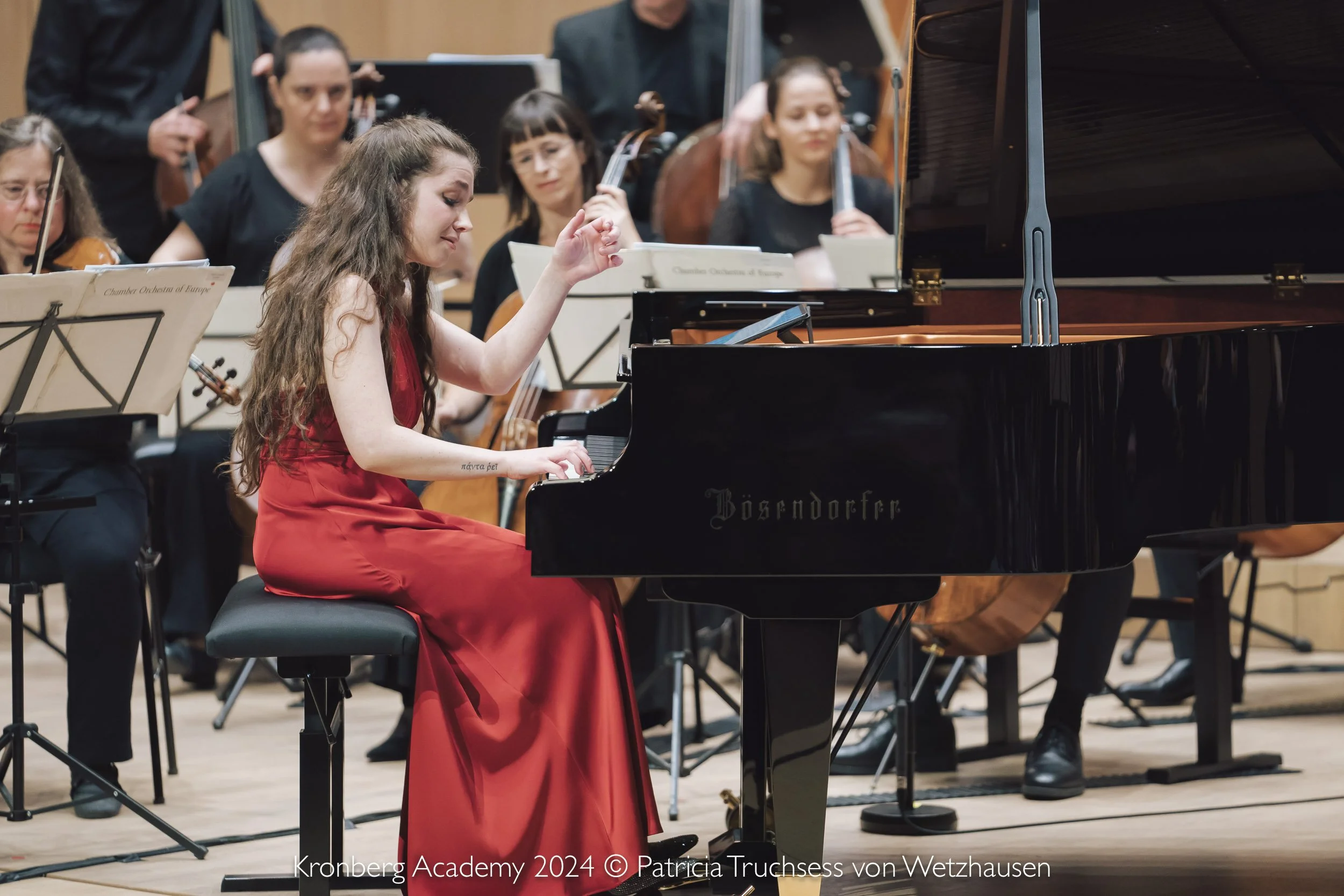 Una giovane donna in abito rosso suona il pianoforte durante un concerto con un'orchestra di musicisti in abiti neri sul palco.