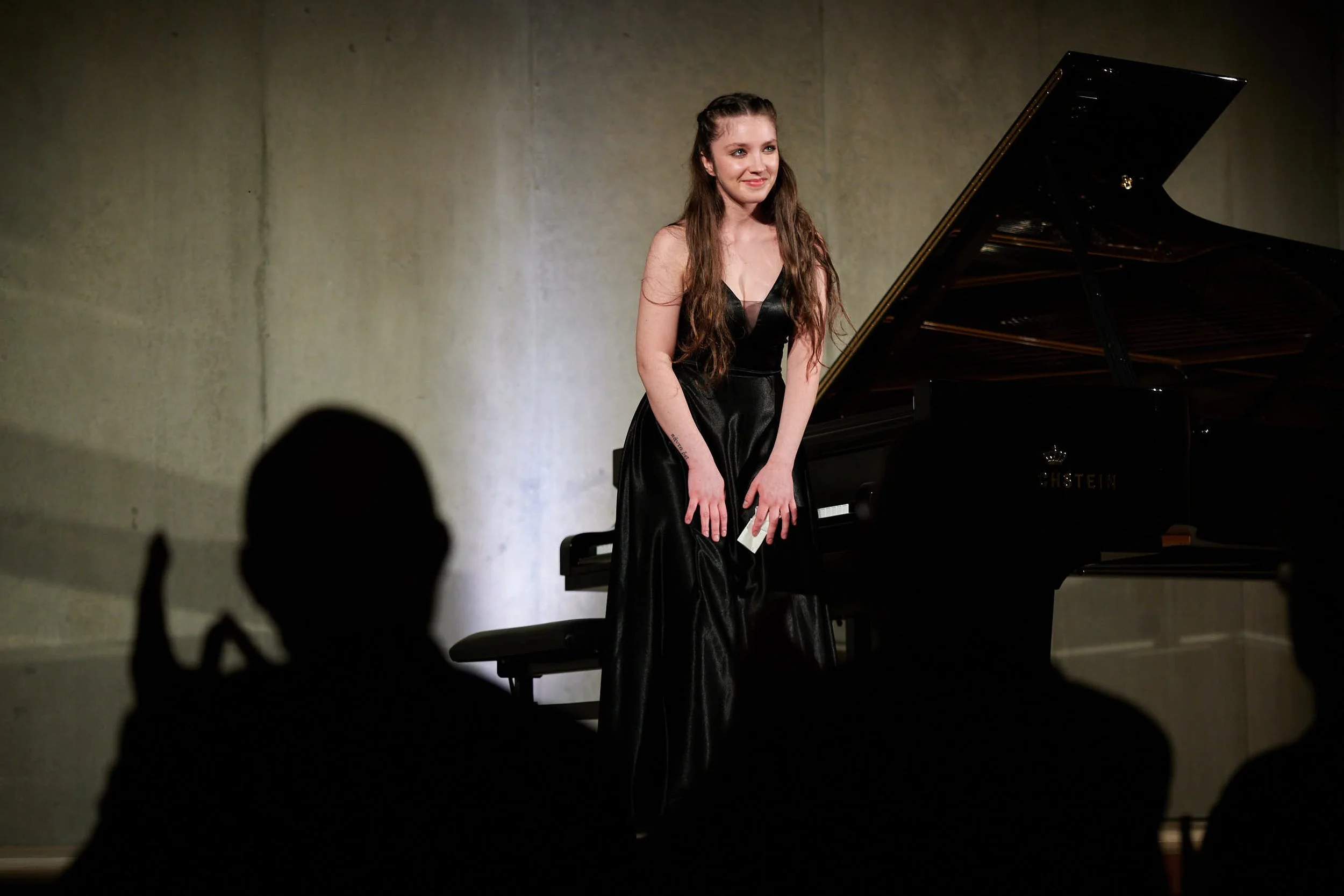 Una giovane donna in abito nero si inchina sul palco di fronte a un pubblico, con un pianoforte a coda dietro di lei, durante un concerto.