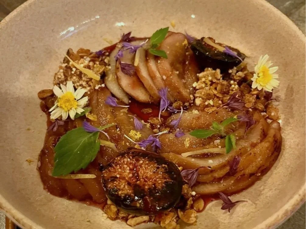 A plated dish with roasted eggplant, sliced pears, edible flowers, crushed nuts, and green herb leaves, garnished with purple edible flowers.