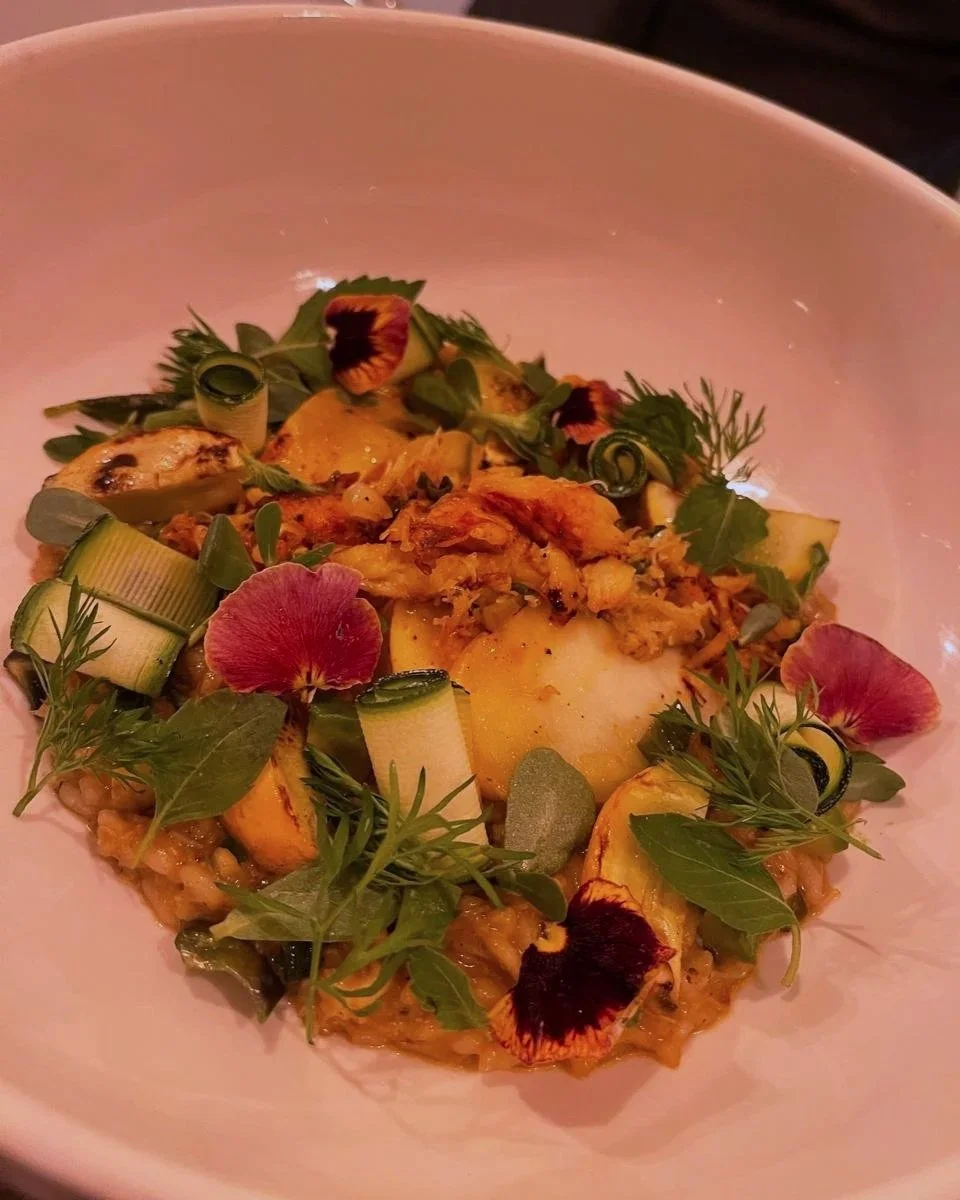 A plated gourmet dish with sliced yellow vegetables, green herbs, edible flowers, and a creamy sauce served on a white plate.