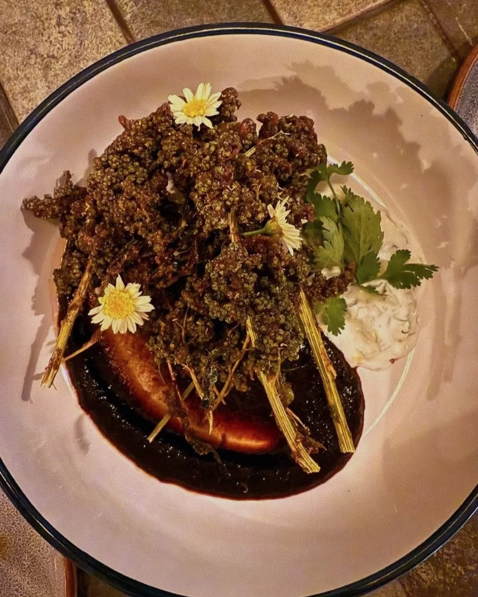 Close-up of a plated dish with cooked broccoli, garnished with flowers and cilantro, served with a side of creamy sauce and a slice of tomato under the vegetables.