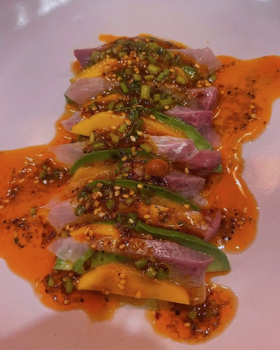 Sliced raw fish topped with a spicy chili sauce, chopped green onions, and sesame seeds, laid out on a white surface.