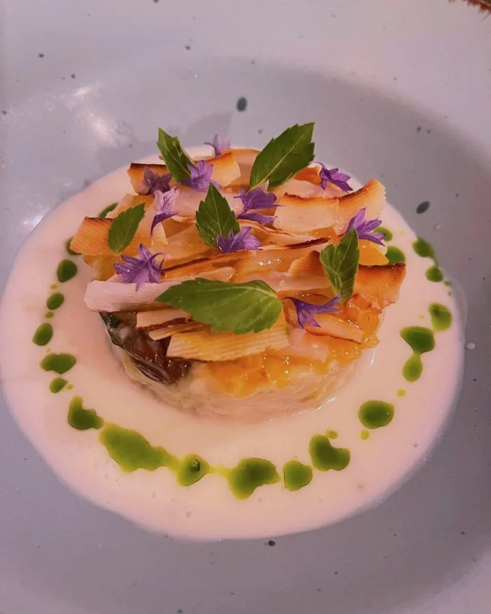 A plated gourmet dish with a creamy sauce, topped with toasted almond slices, purple edible flowers, and green herbs, garnished with a drizzle of green sauce.