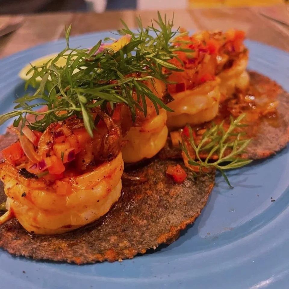Close-up of a gourmet dish featuring shrimp on a blue plate, garnished with fresh herbs and topped with diced red peppers, served on a dark cracker or flatbread.