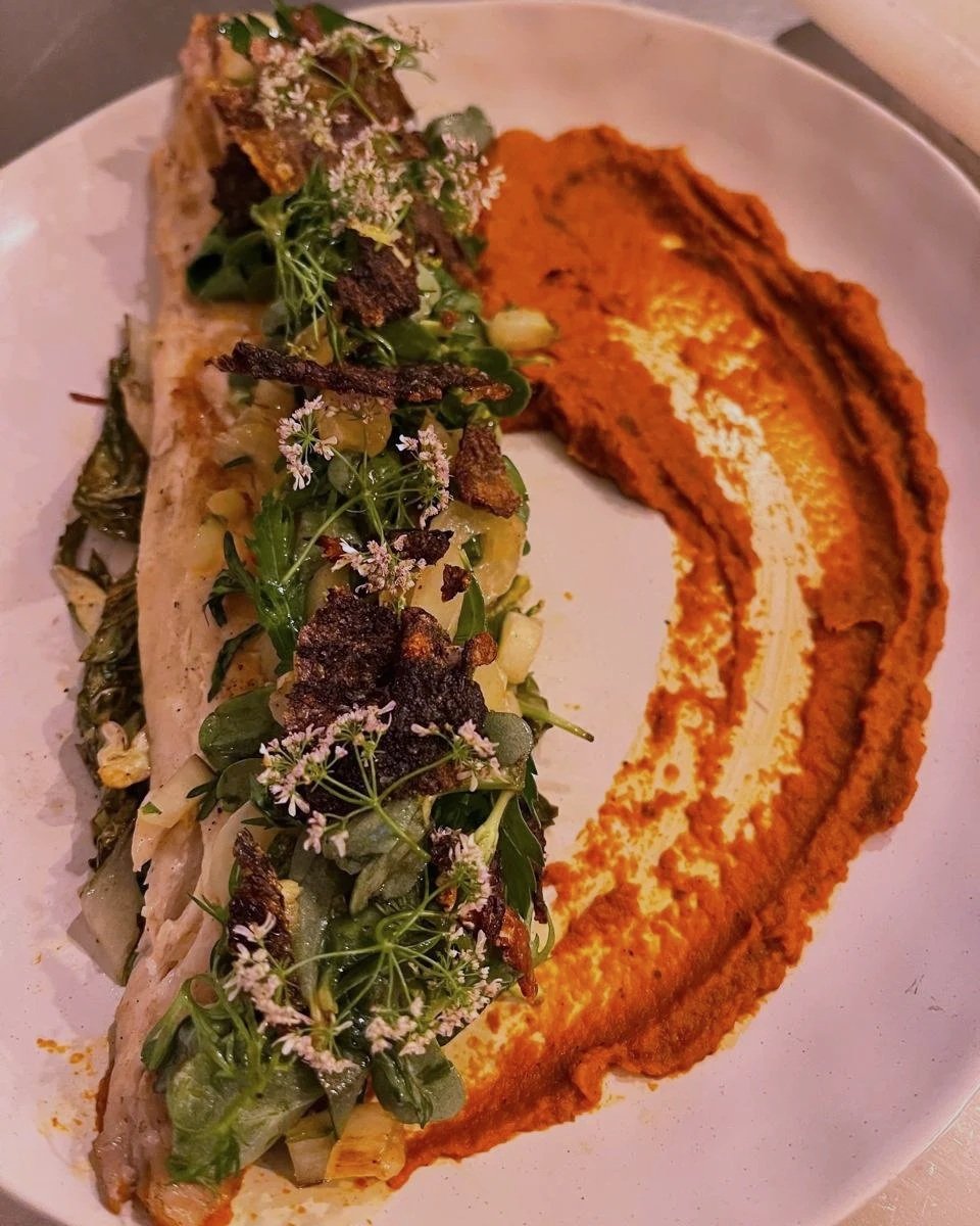 A plate of food with a taco topped with greens, edible flowers, and crispy elements, served alongside a smear of orange-colored sauce or mash.