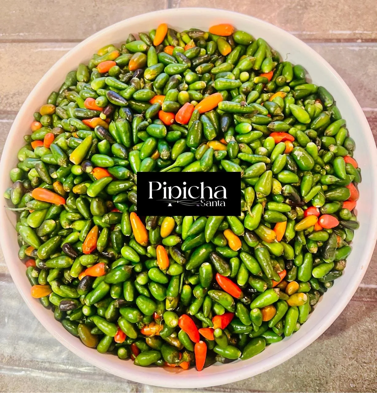 A white bowl filled with small, green, and orange chili peppers on a tiled surface with a label that says "Pipicha Santa" in the center.