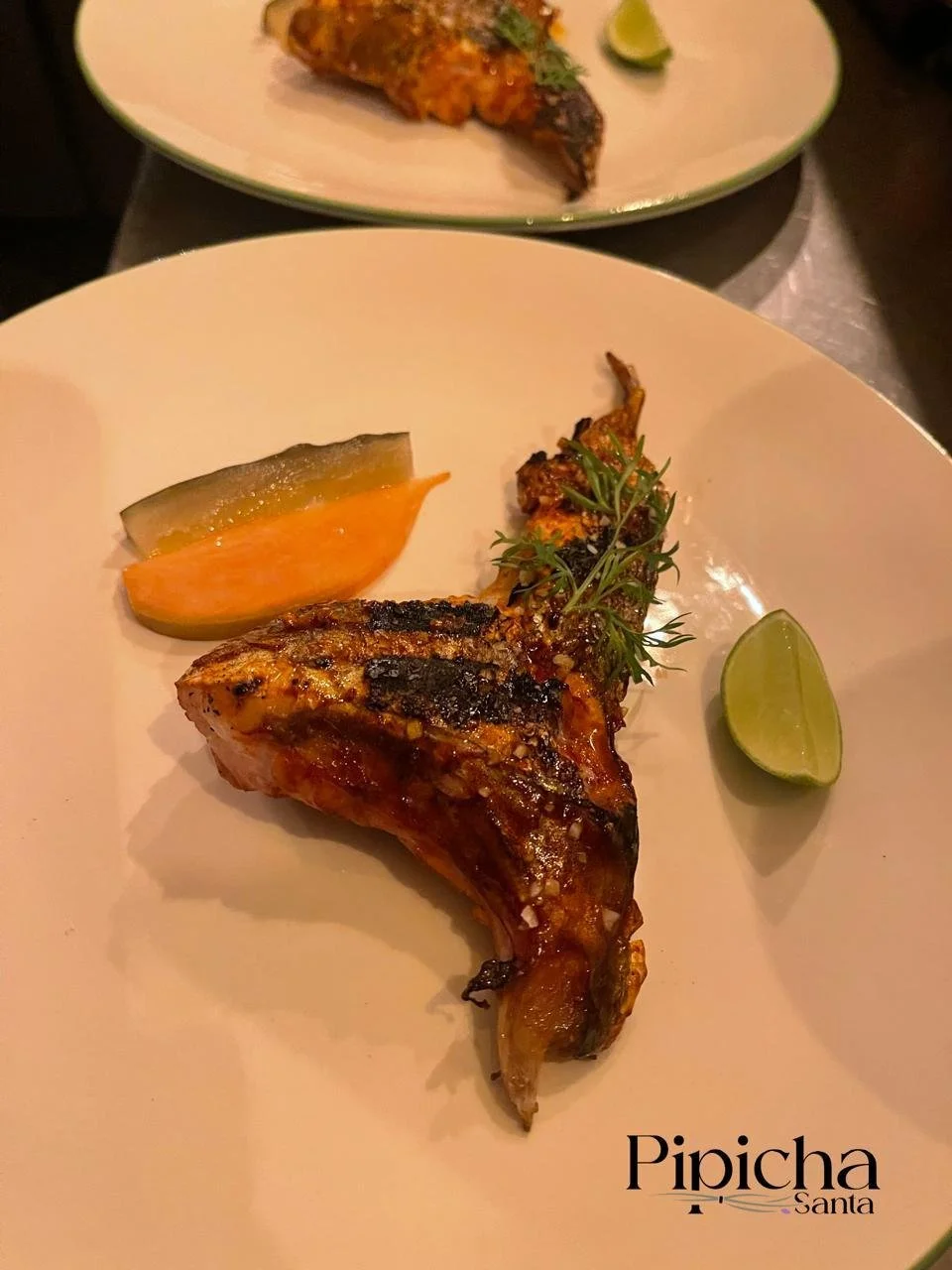 Grilled chicken leg quarter with lime wedge, pickle slice, and herb garnish on a white plate, with another plate of chicken in the background.