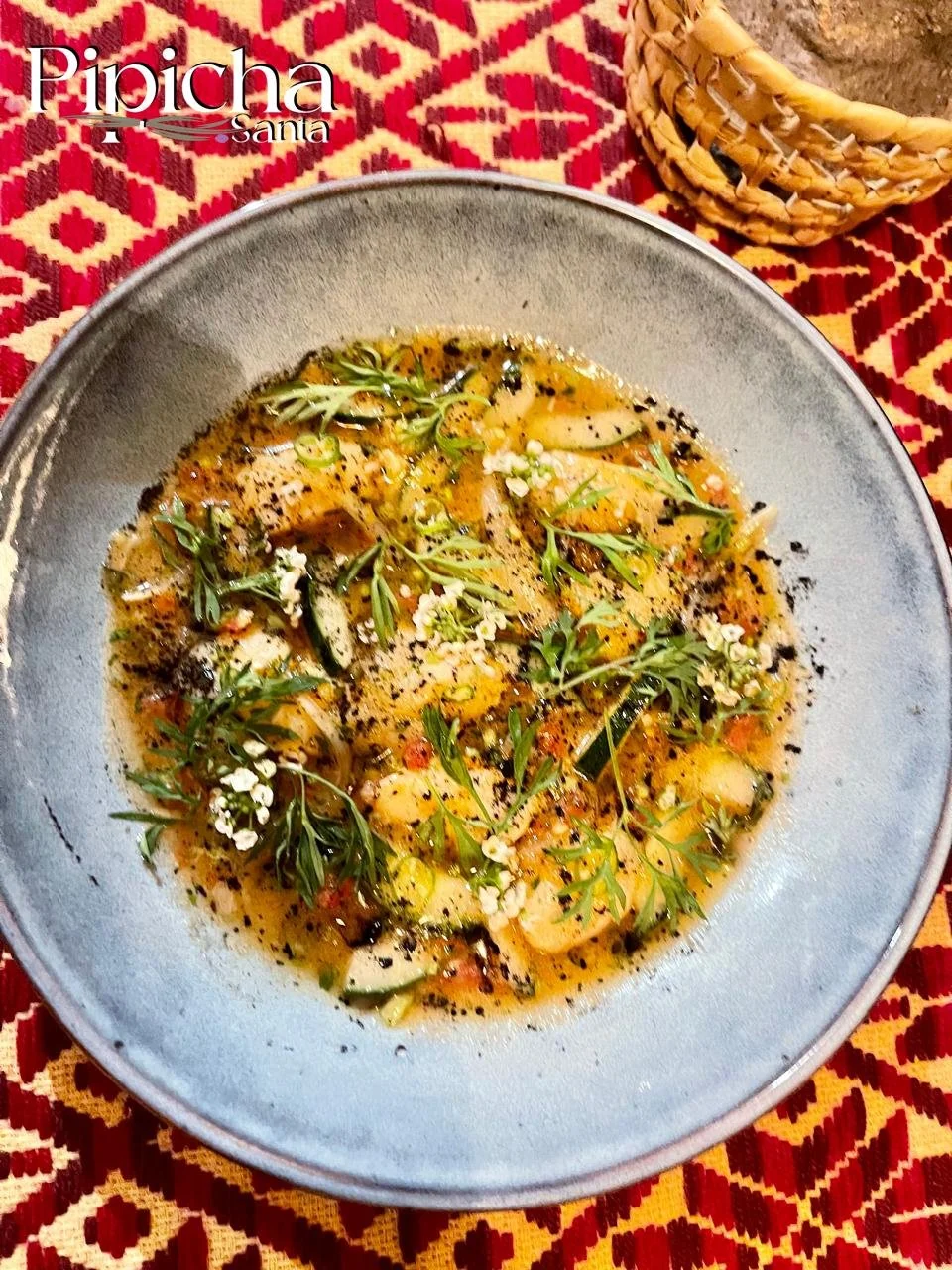 A bowl of traditional Mexican dish, possibly Sopa Tarasca, featuring a broth with herbs, small vegetables, and garnished with fresh herbs and microgreens, served on a decorative tablecloth.