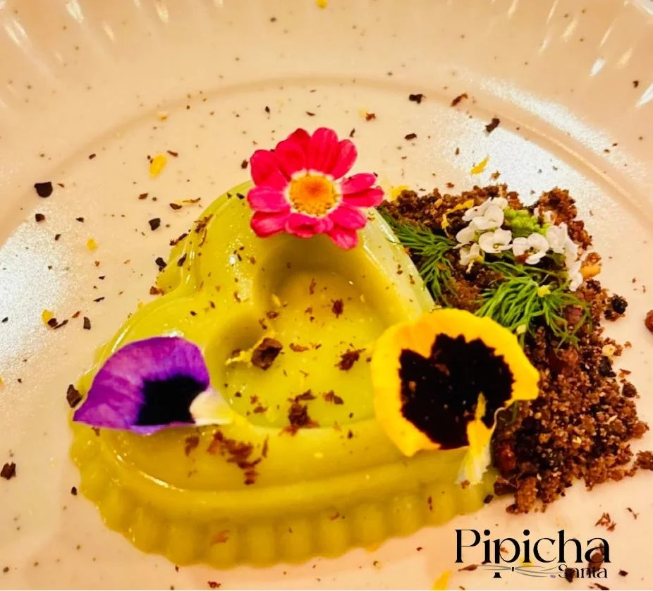 A dessert with a yellow-green topping, garnished with edible flowers, herbs, and chocolate shavings.