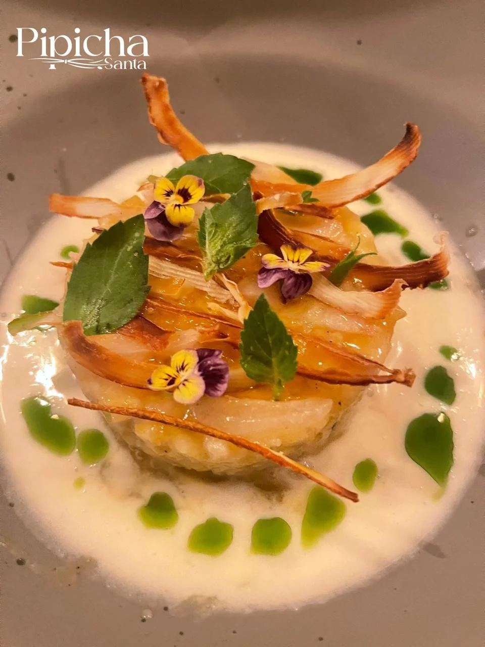 Elegant dish with roasted vegetables, edible flowers, fresh herbs, served in a light creamy sauce with green oil drops, in a gray bowl.