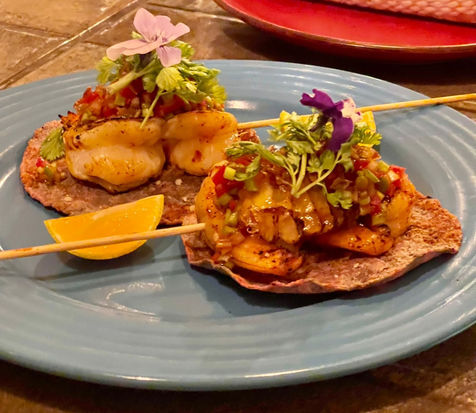 Two seafood tostadas topped with diced vegetables and microgreens, served on a blue plate with a lemon wedge.