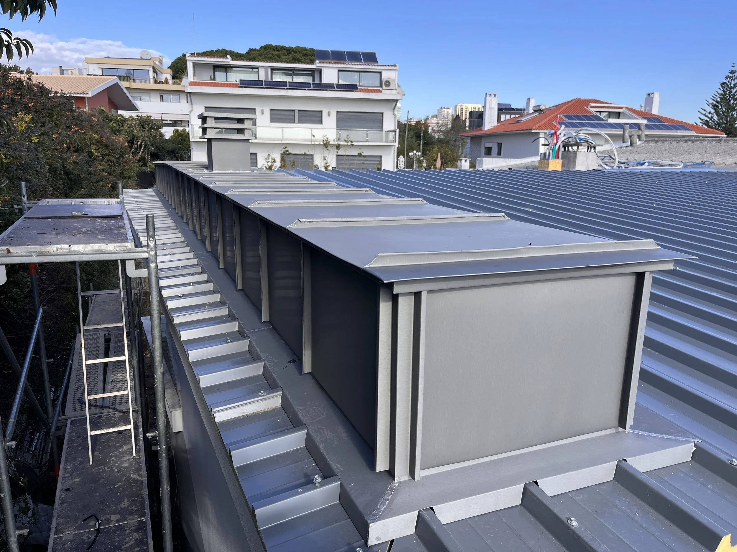 Imagem mostra uma instalação de aberturas de ventilação em uma cobertura de telas metálicas, com casas residenciais ao fundo, incluindo painéis solares.