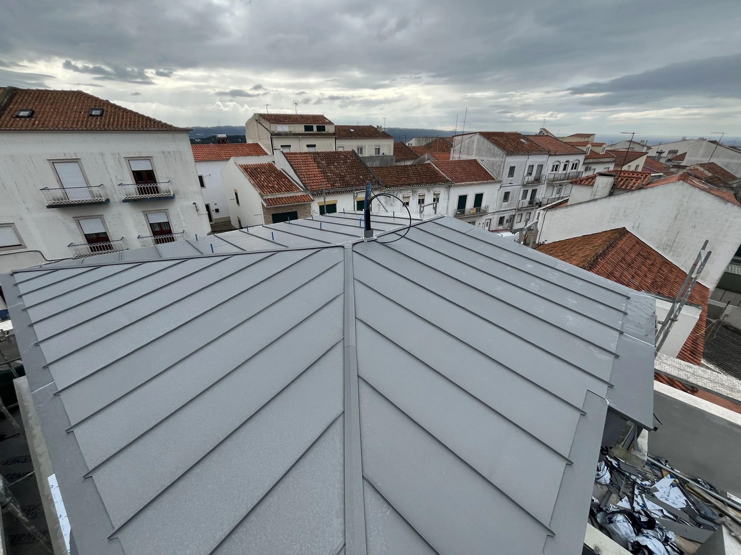 Vista do telhado de uma construção com cobertura de metal na cidade, com casas de telhado de cerâmica vermelha ao fundo e céu nublado.