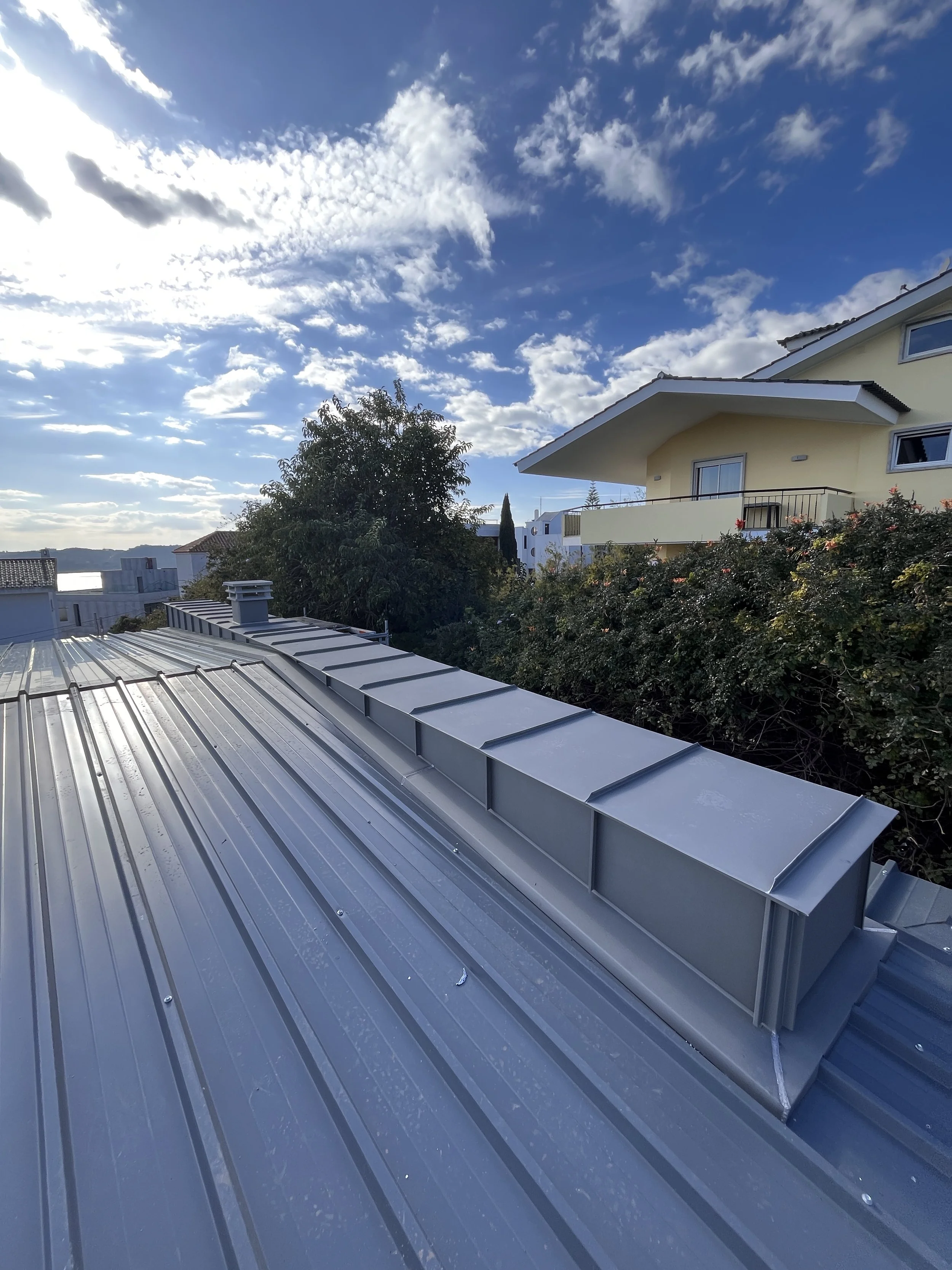 Telhado de metal prateado em primeiro plano, com uma chaminé, sob céu parcialmente nublado, com árvores e casas ao fundo.