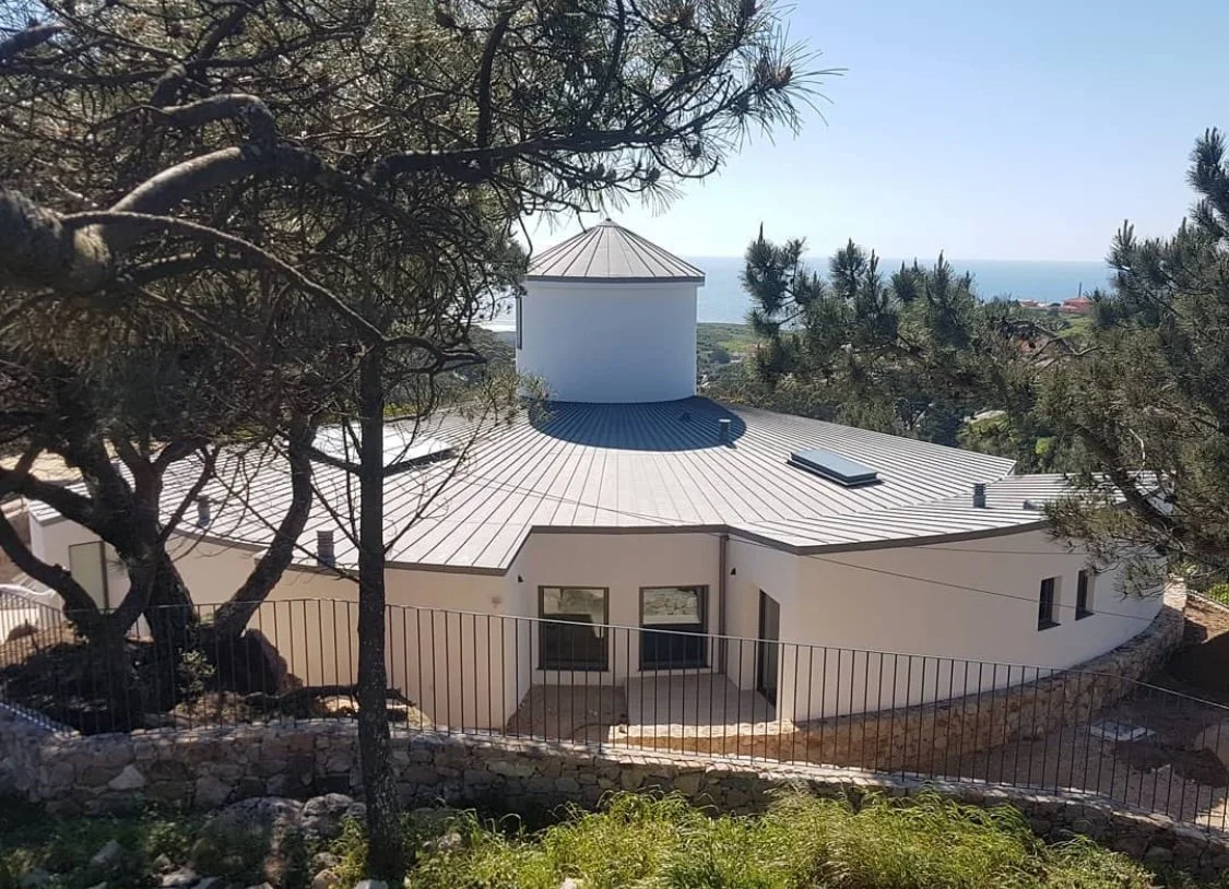 Casa moderna com telhado de metal, rodeada de árvores, vista para o mar ao fundo.