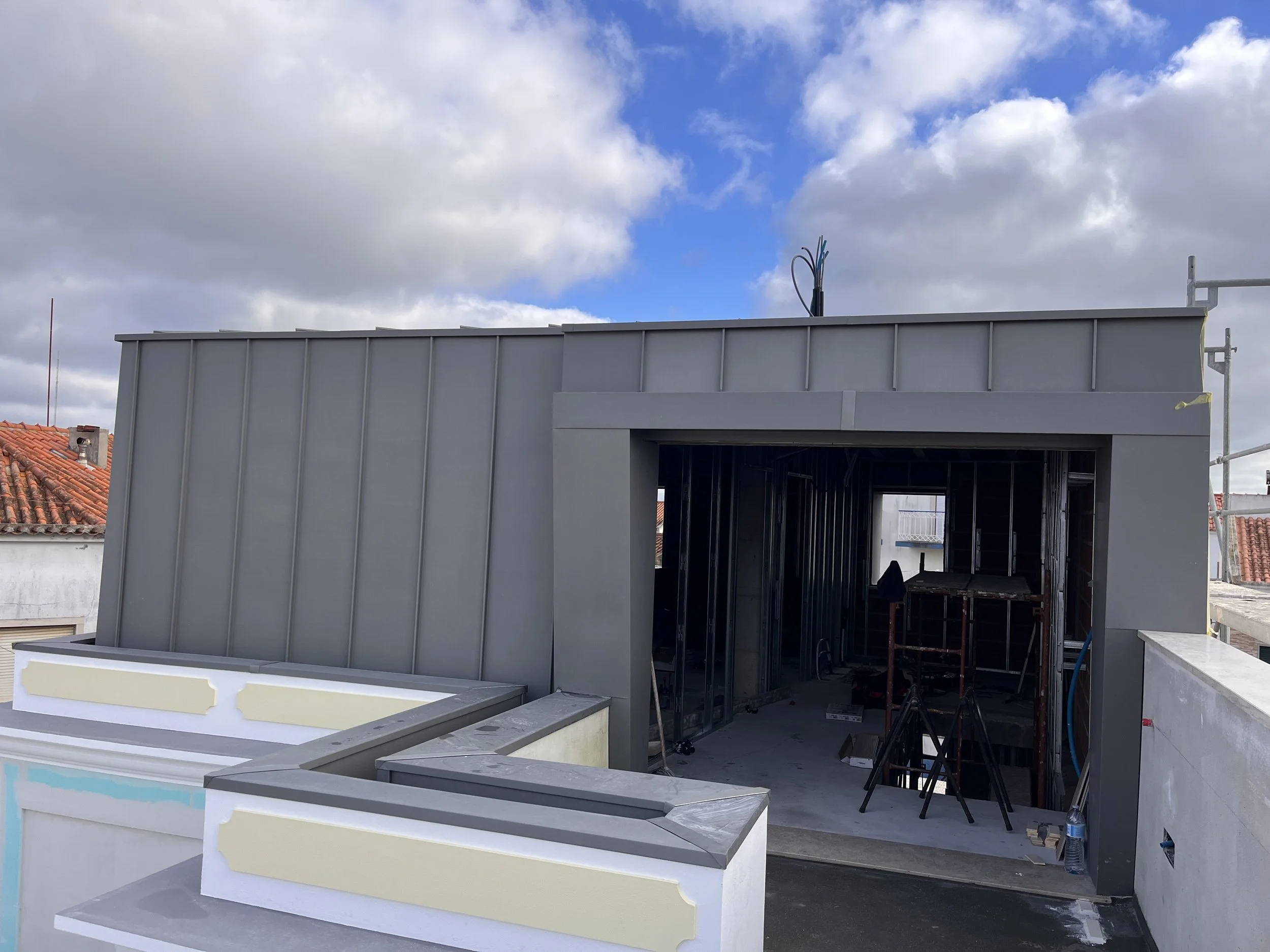 Construção de uma estrutura de edifício com paredes de metal cinza, sob céu parcialmente nublado, próximo a telhados de barro vermelho.