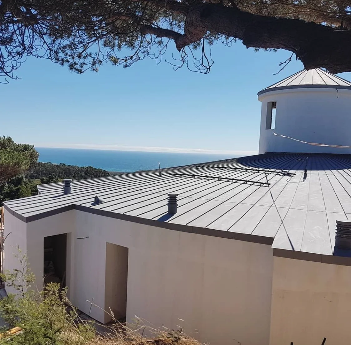 Casa moderna com telhado de metal na encosta com vista para o mar e céu claro ao fundo, árvores ao redor.