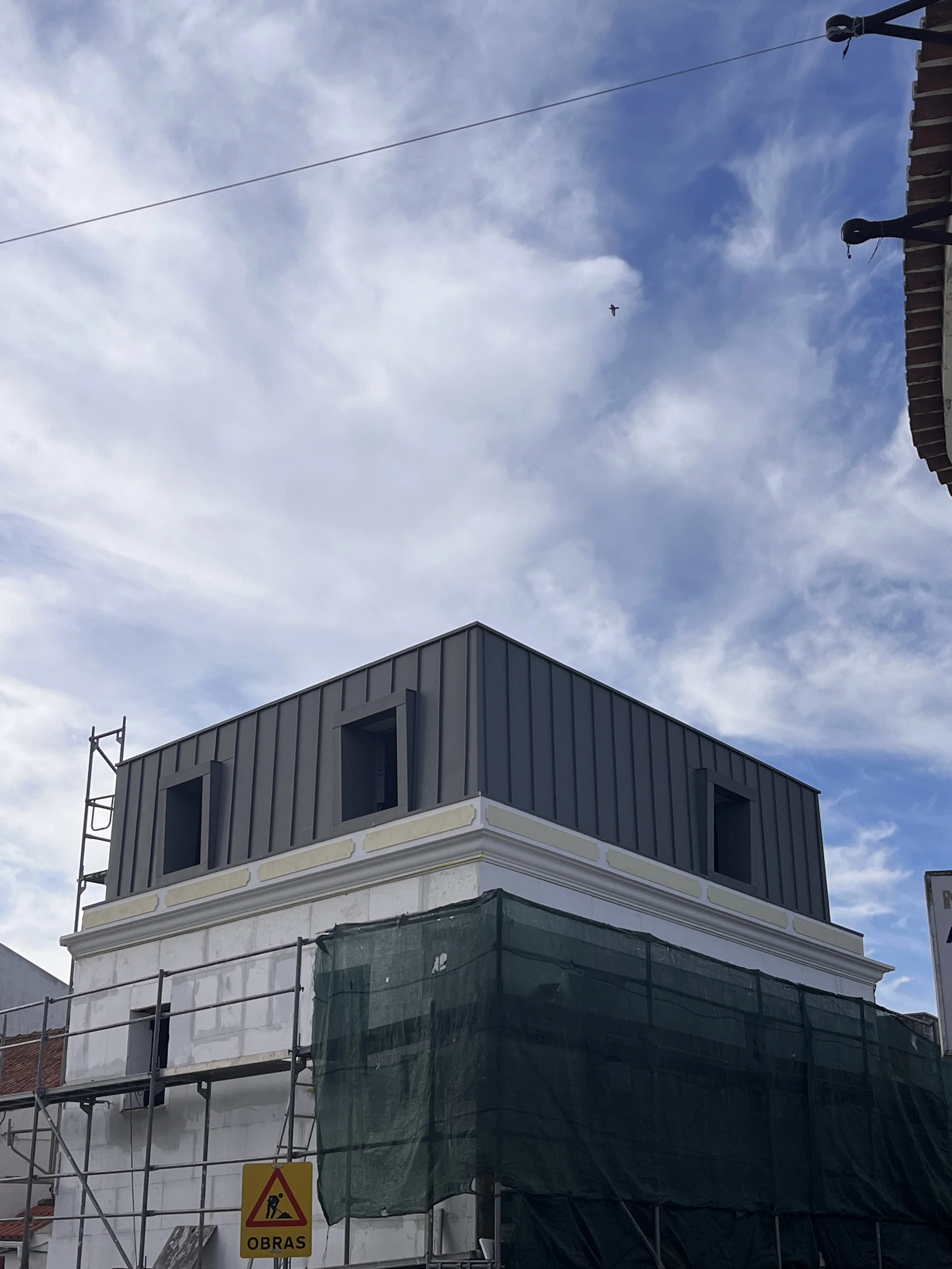 Construção de um edifício com duas janelas abertas na parte superior, cercado por andaimes, com céu azul e nuvens ao fundo.