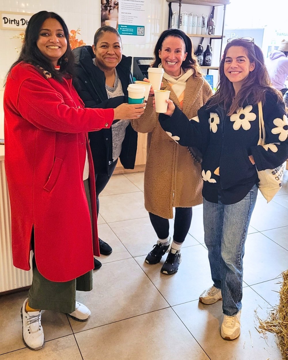Four women inside a coffee shop, holding coffee cups and smiling at the camera. They are dressed casually, with one in a red coat, one in a black jacket, one in a tan coat, and one in a black coat with white flower patterns. The background shows coff