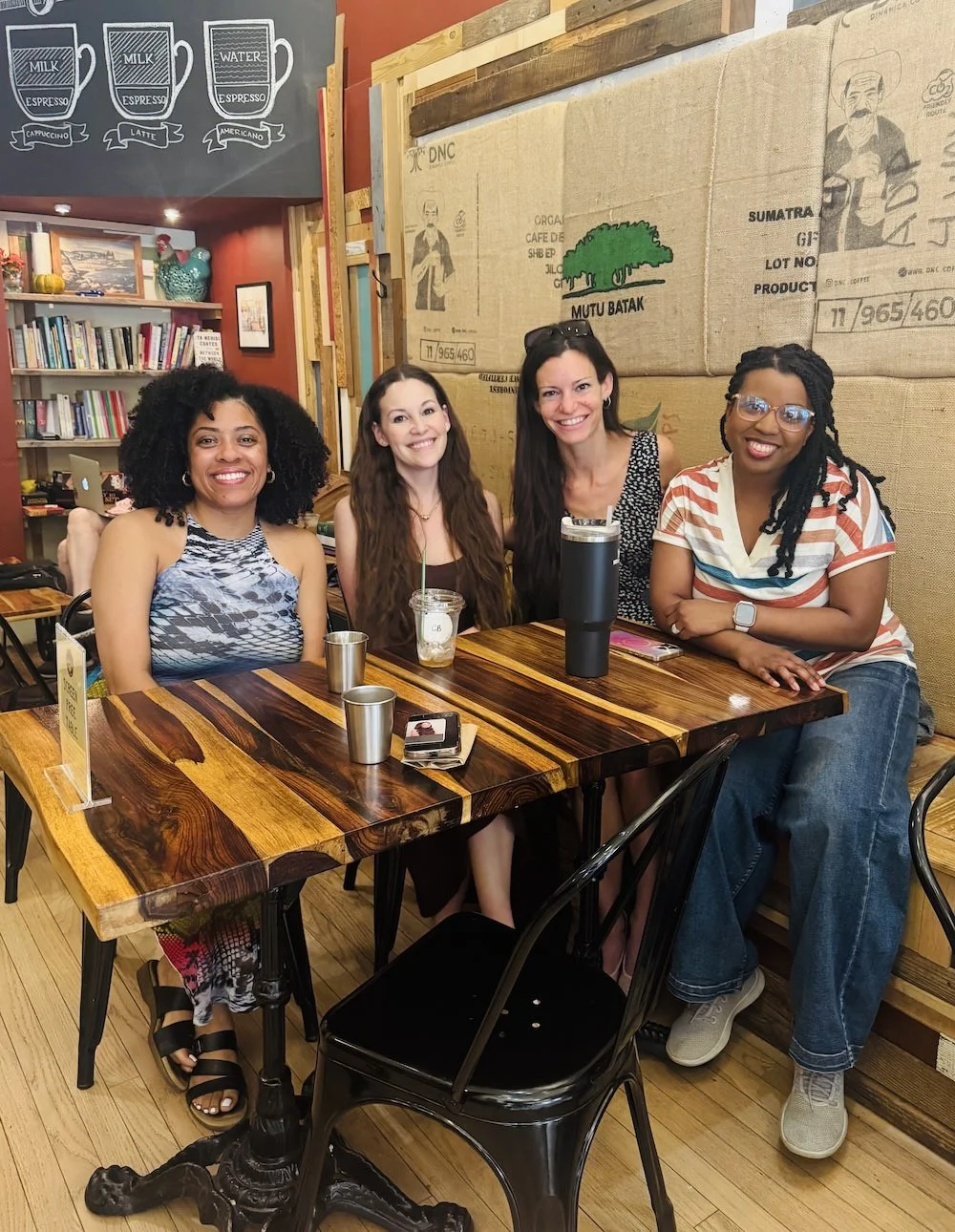 Four women sitting at a wooden table inside a Java Love Coffee Roasters in Montclair, NJ, smiling at the camera. Behind them are bookshelves and a mural with coffee-related drawings and words.