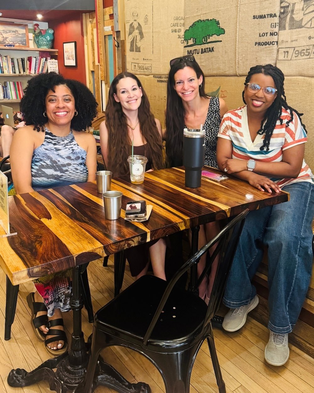 Four women sitting at a wooden table in a cozy cafe, smiling at the camera.