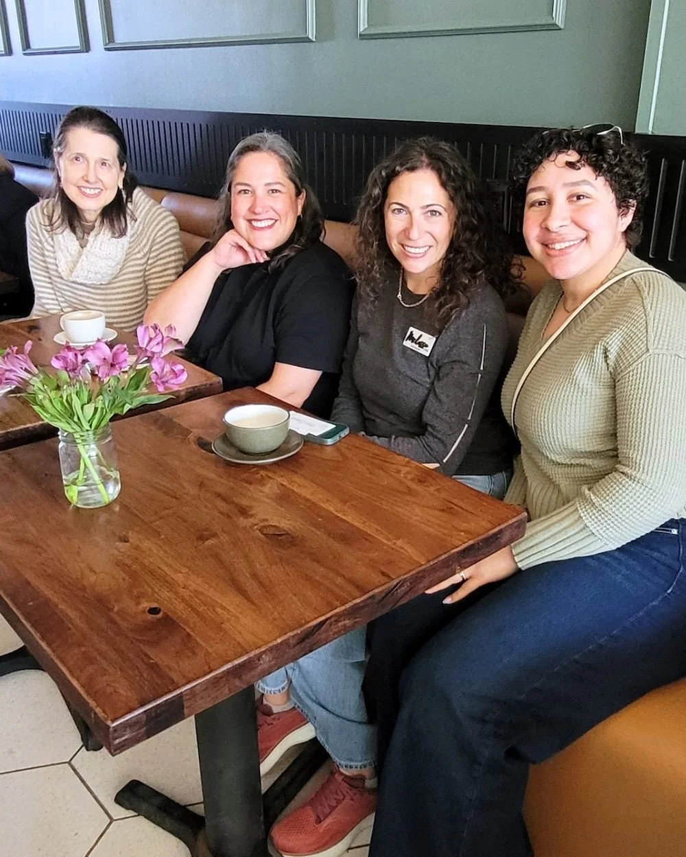 Four women entrepreneurs sitting at a wooden table in a restaurant, with cups of coffee or tea, chatting during a coffee meetup in New Jersey.