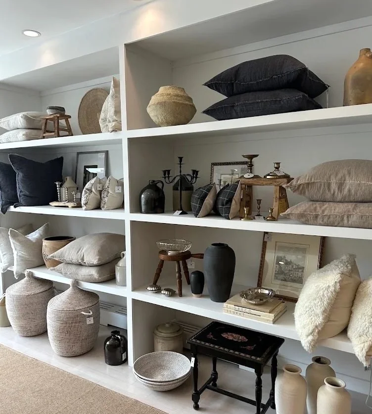 Interior display at GATHER in Millburn, NJ, featuring vintage furniture, decorative pillows, ceramics, antiques, and curated home décor on white shelving.