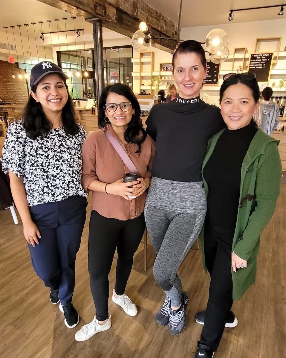Four women standing together inside a coffee shop, smiling at the camera.