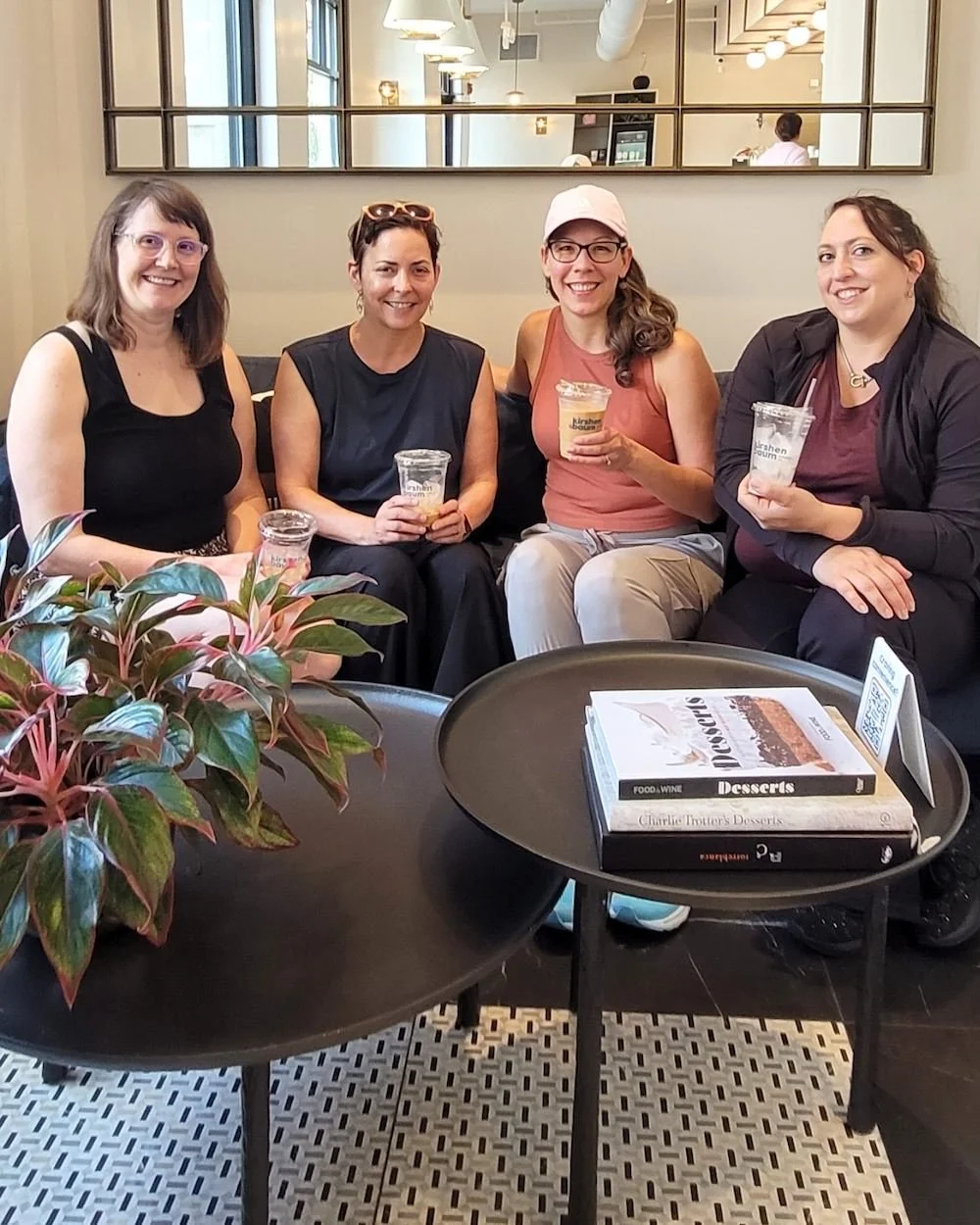 Four women sitting together on a couch in a Morristown, NJ cafe, smiling and holding drinks, with books on a tray table in front. The background shows a mirror and cafe interior.