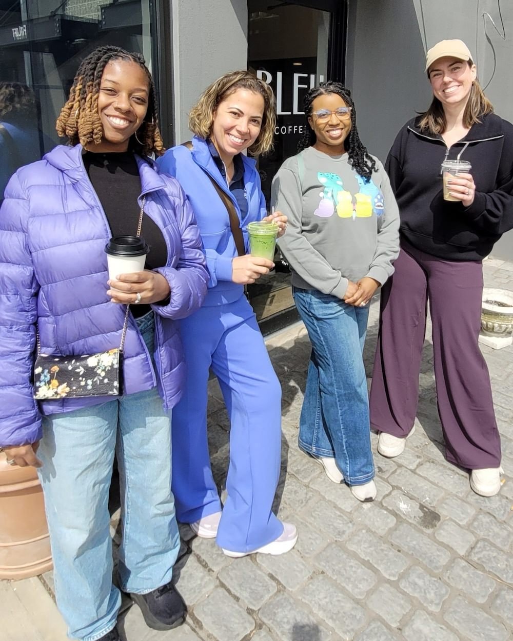 Four women standing outside a coffee shop in Montclair, smiling, holding drinks, dressed casually and warmly, with a black storefront and gray sidewalk behind them.