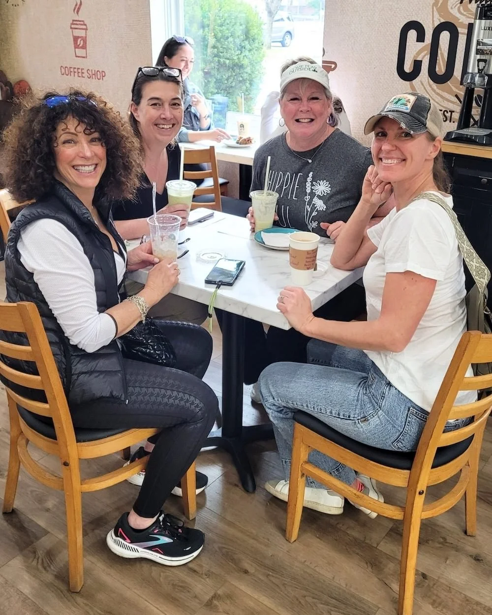 Four women business owners sitting at a table in a coffee shop, smiling and enjoying drinks, with a fifth woman in the background at another table.