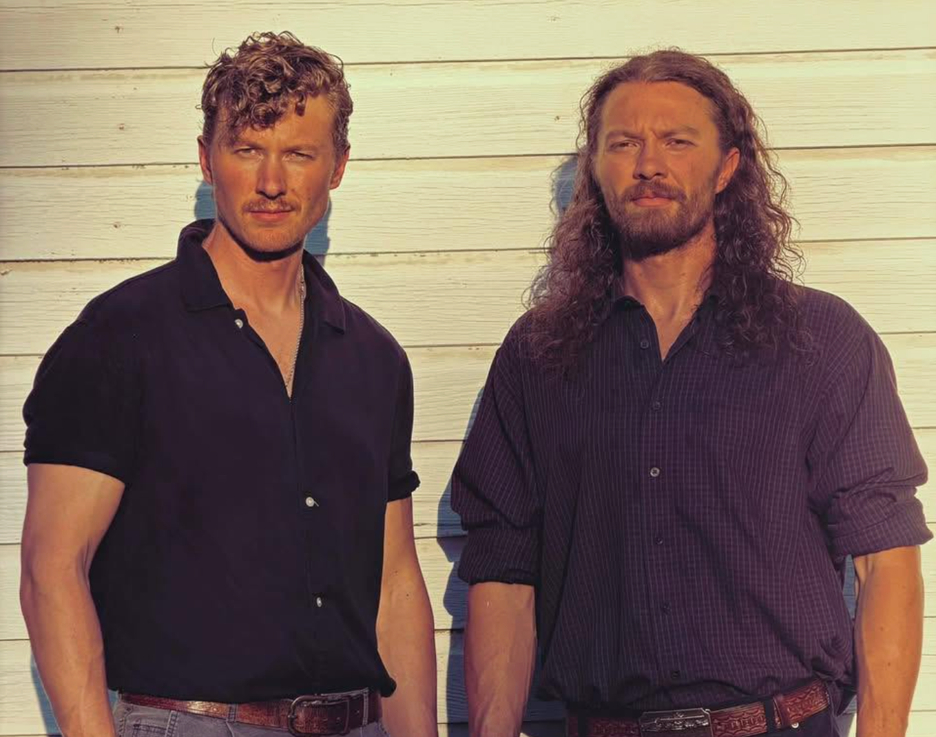 Two men outdoors with musical instruments, holding cups with 'No Proof' slogans, smiling in front of a wooden fence and trees.