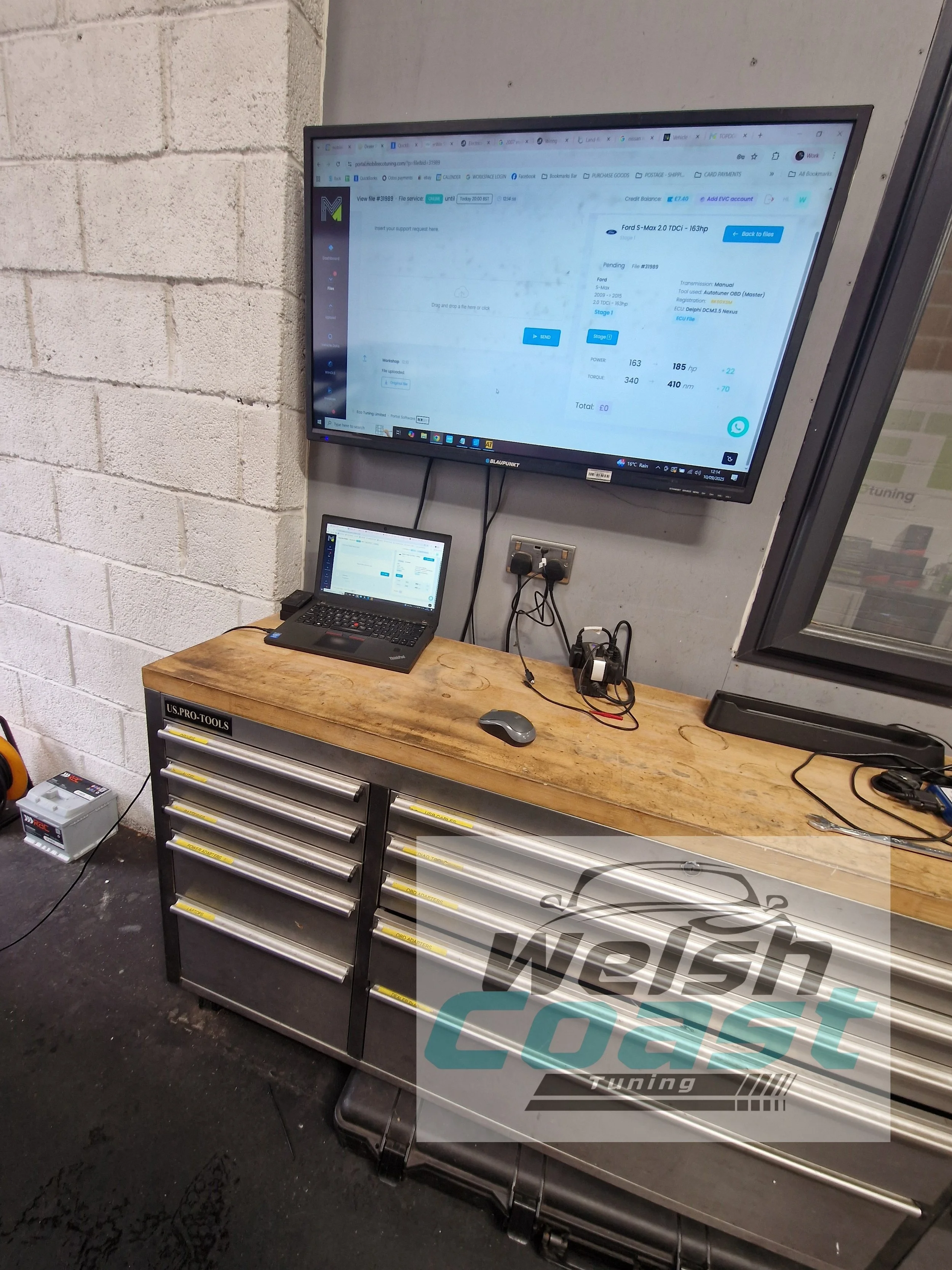 A workshop with a metal tool chest, a wooden work surface, and a mounted monitor displaying a car tuning software. A laptop and a computer mouse are on the work surface, with cables connected to the monitor and power outlets.